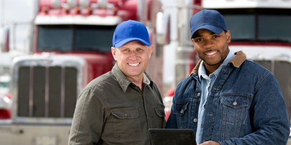 Two male fleet drivers smiling while holding a tablet in front of a large red commercial truck.